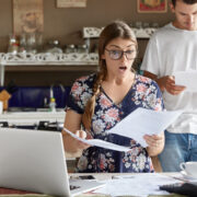 Young female looks with shocked expression into papers, recieve high taxes and had to pay much money for bills, sits against kitchen interior, her husband stands behinds, studies documents attentively